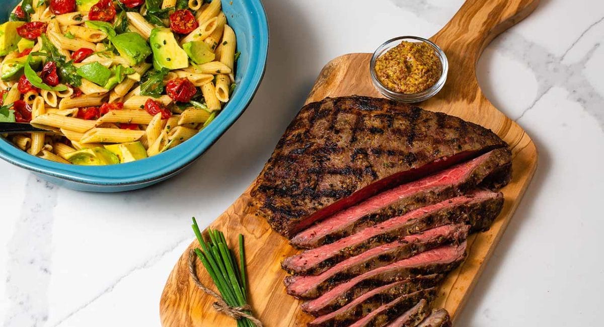 flank steak on a cutting board with a bowl of pasta salad