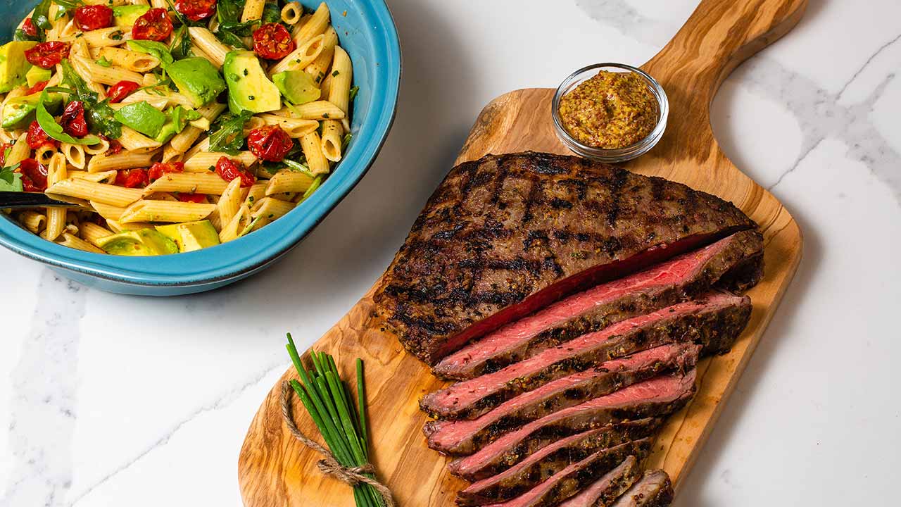 flank steak on a cutting board with a bowl of pasta salad