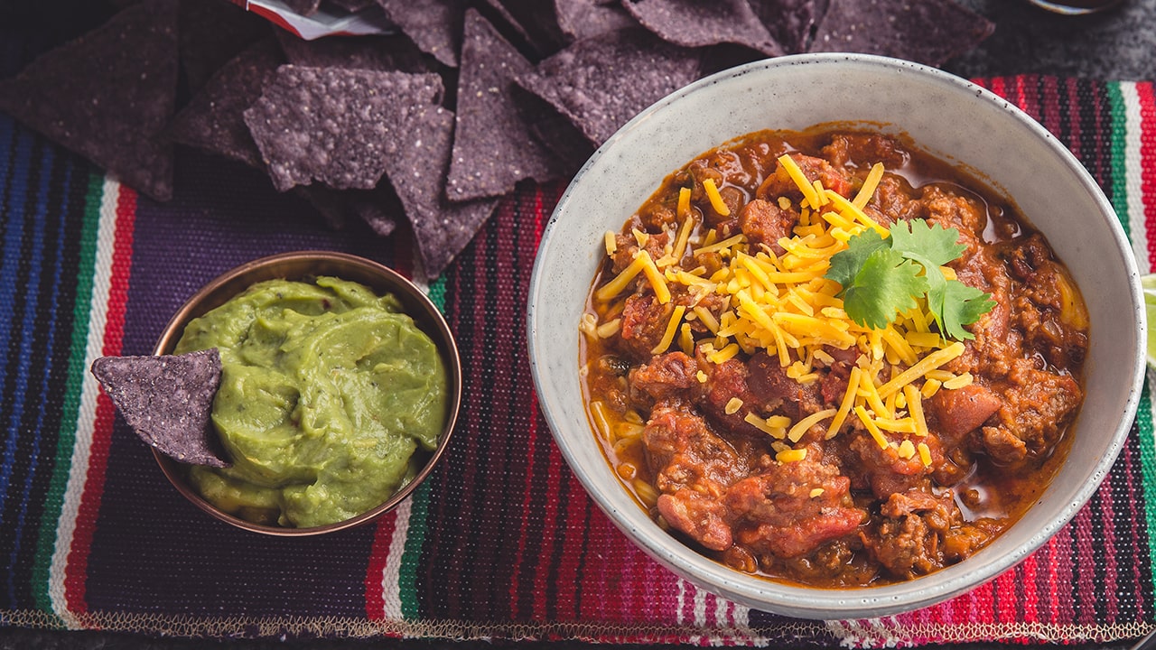 Beef and kidney bean chili in a bowl next to tortilla chips and guacamole