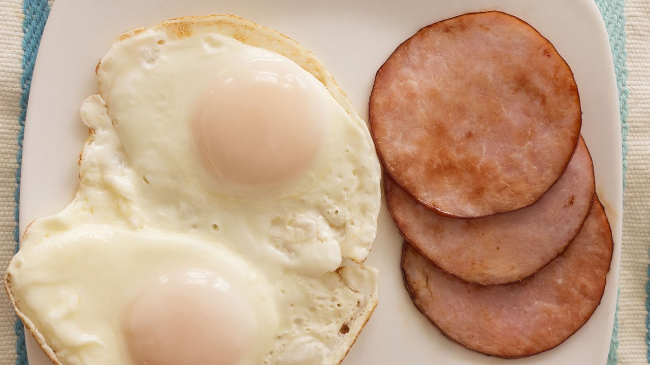 A plate of two eggs, over easy and slices of Canadian bacon on a pastel tablecloth.