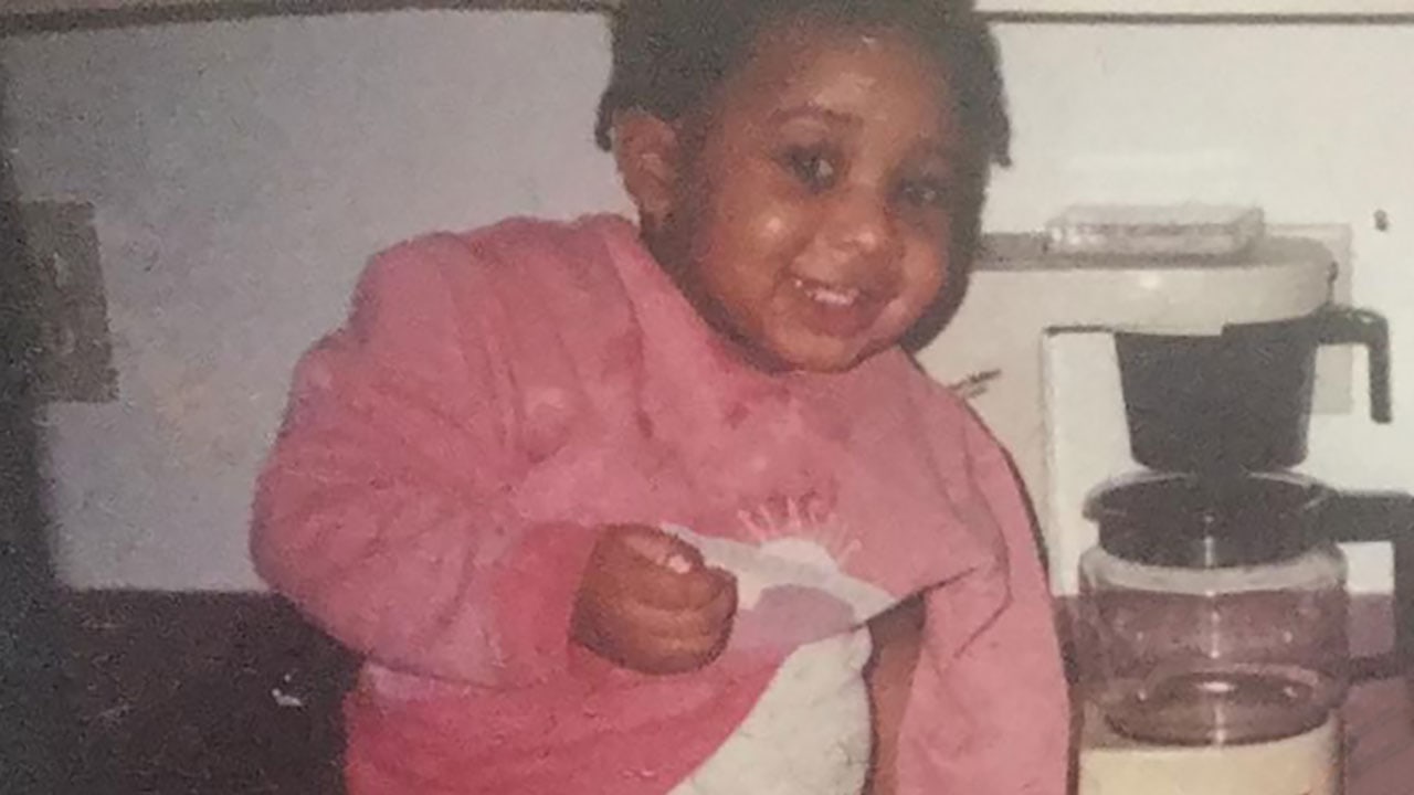 Smiling child in a sweatshirt sitting on a kitchen countertop with a coffee maker in the background.