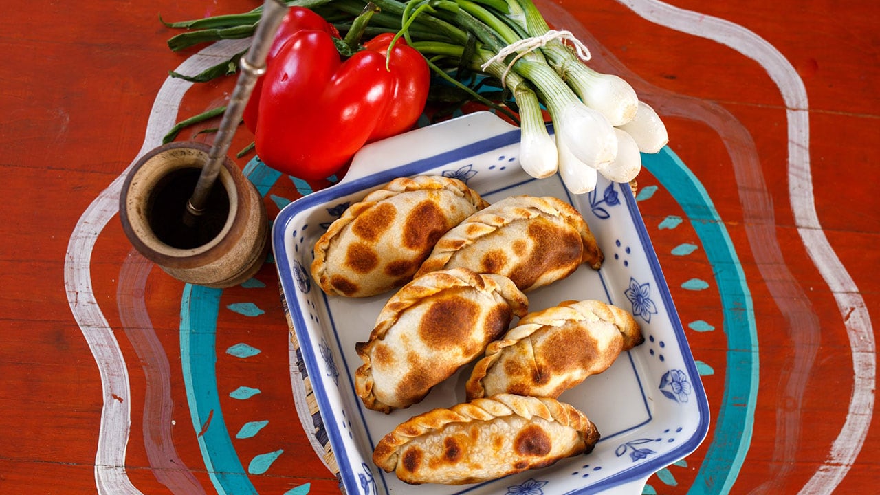 empanadas in a casserole dish next to a jar of sauce, red pepper, and bunch of scallions