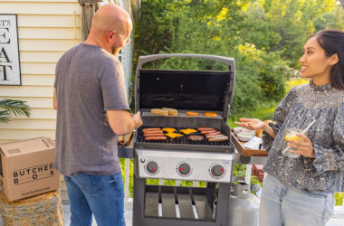 Two people with drinks standing by the grill as it cooks meat