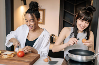 two people cooking in a kitchen: one is cracking an egg and the other is slicing cheese.