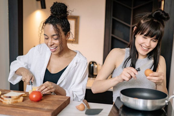 two people cooking in a kitchen: one is cracking an egg and the other is slicing cheese.