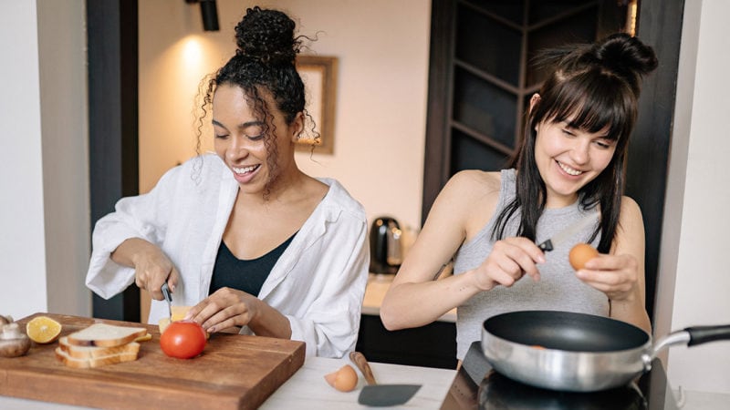 two people cooking in a kitchen: one is cracking an egg and the other is slicing cheese.
