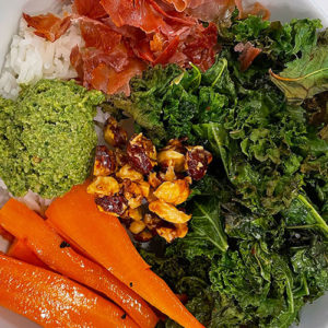 rice topped with roasted kale, carrots, proscuitto, hazelnuts, and basil pesto in a bowl