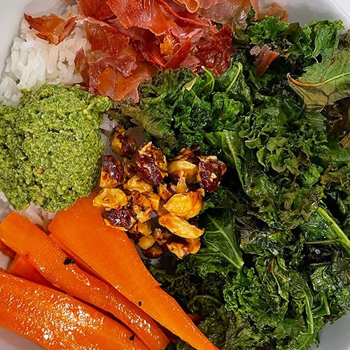 rice topped with roasted kale, carrots, proscuitto, hazelnuts, and basil pesto in a bowl