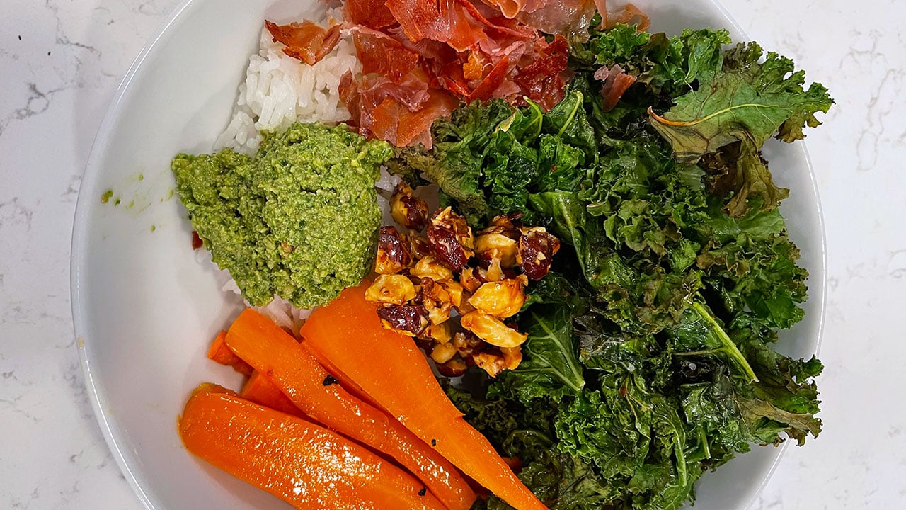 rice topped with roasted kale, carrots, proscuitto, hazelnuts, and basil pesto in a bowl