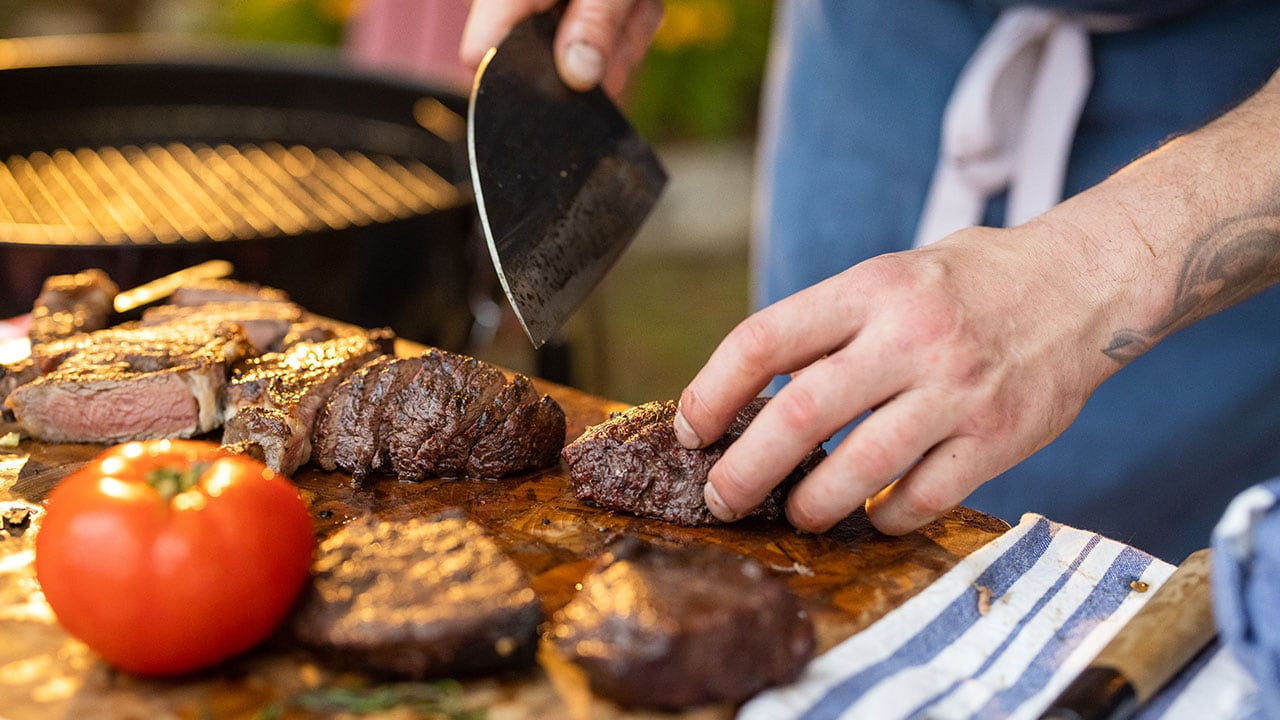 person using a cleaver knife to slice grilled steak on a cutting board