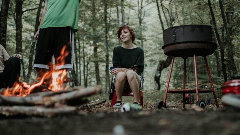 Woman sits by a campfire and a grill in the woods.